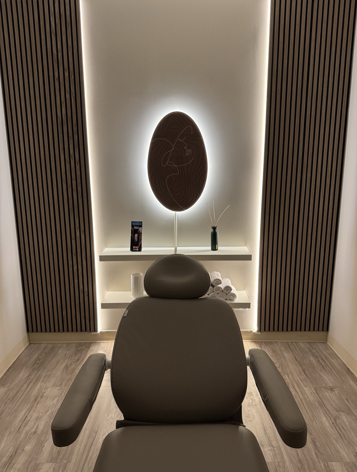 The DK Aesthetics treatment room in Highland Park — warm wood walls, soft backlit line-art, and a single reclining chair.